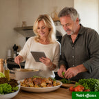 Couple en cuisine suivant une recette sur tablette avec poulet rôti, pommes de terre et légumes frais sur le plan de travail.